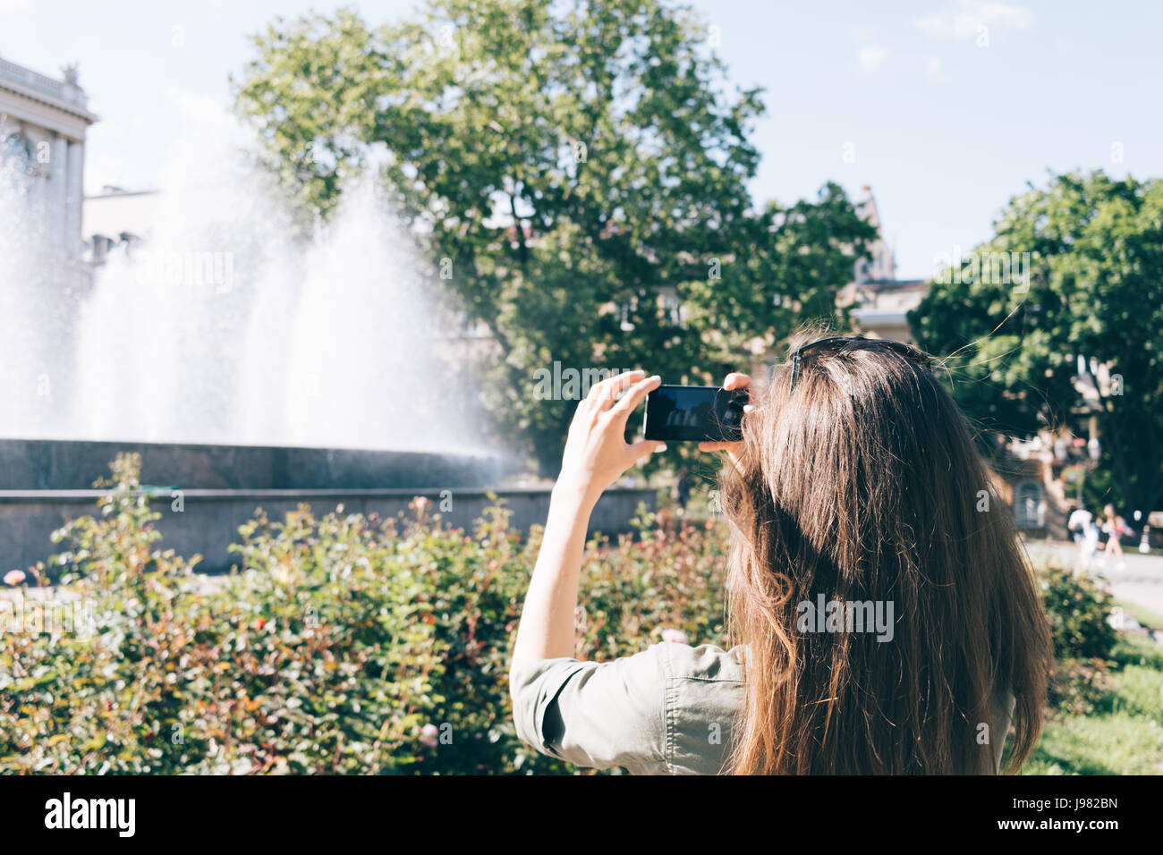 Young woman with long brown hair photographing a fountain in the park ...