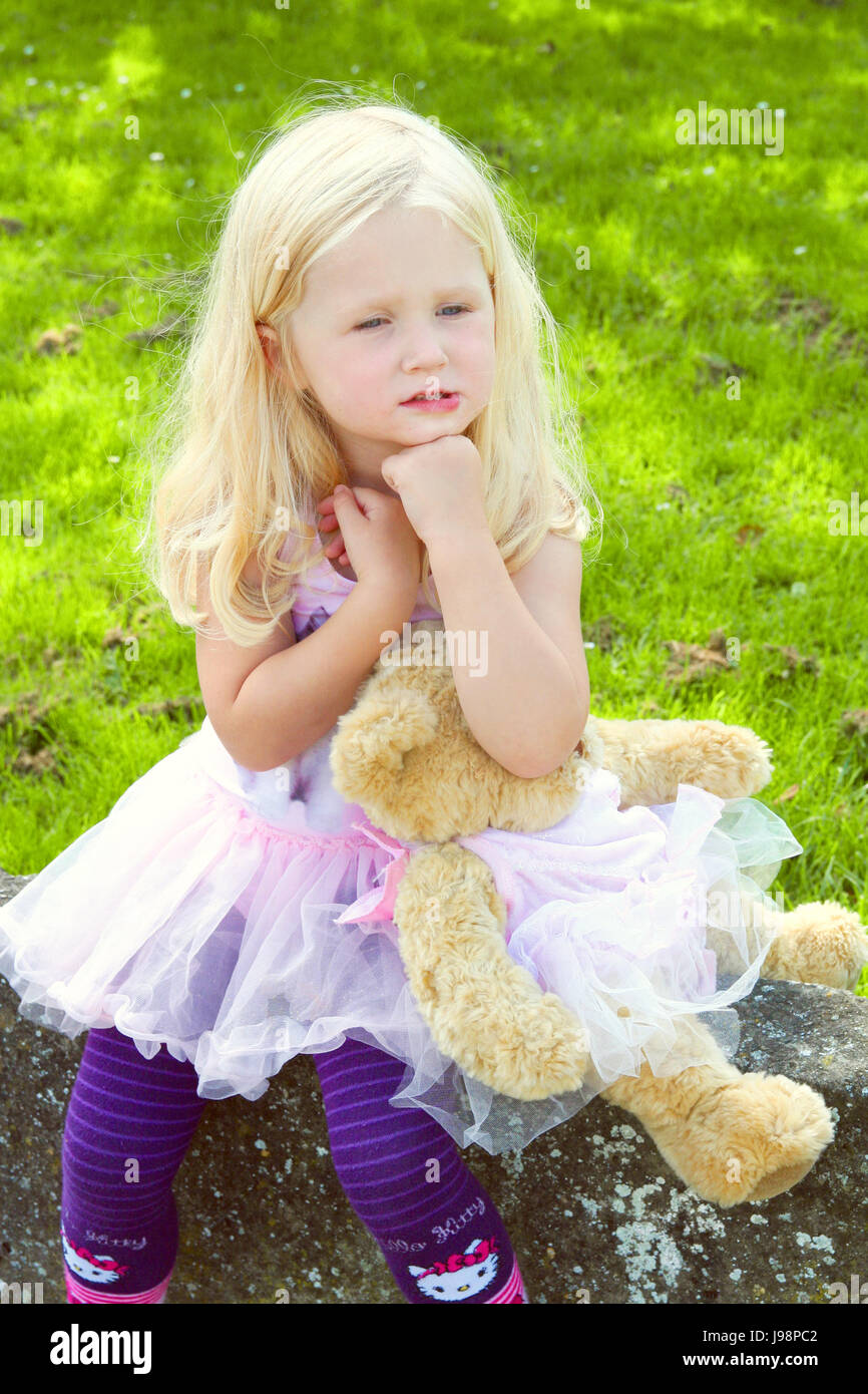 Little three year old girl with long curly blonde hair sitting on the wall child holding her matching teddy bear wearing a tutu Stock Photo