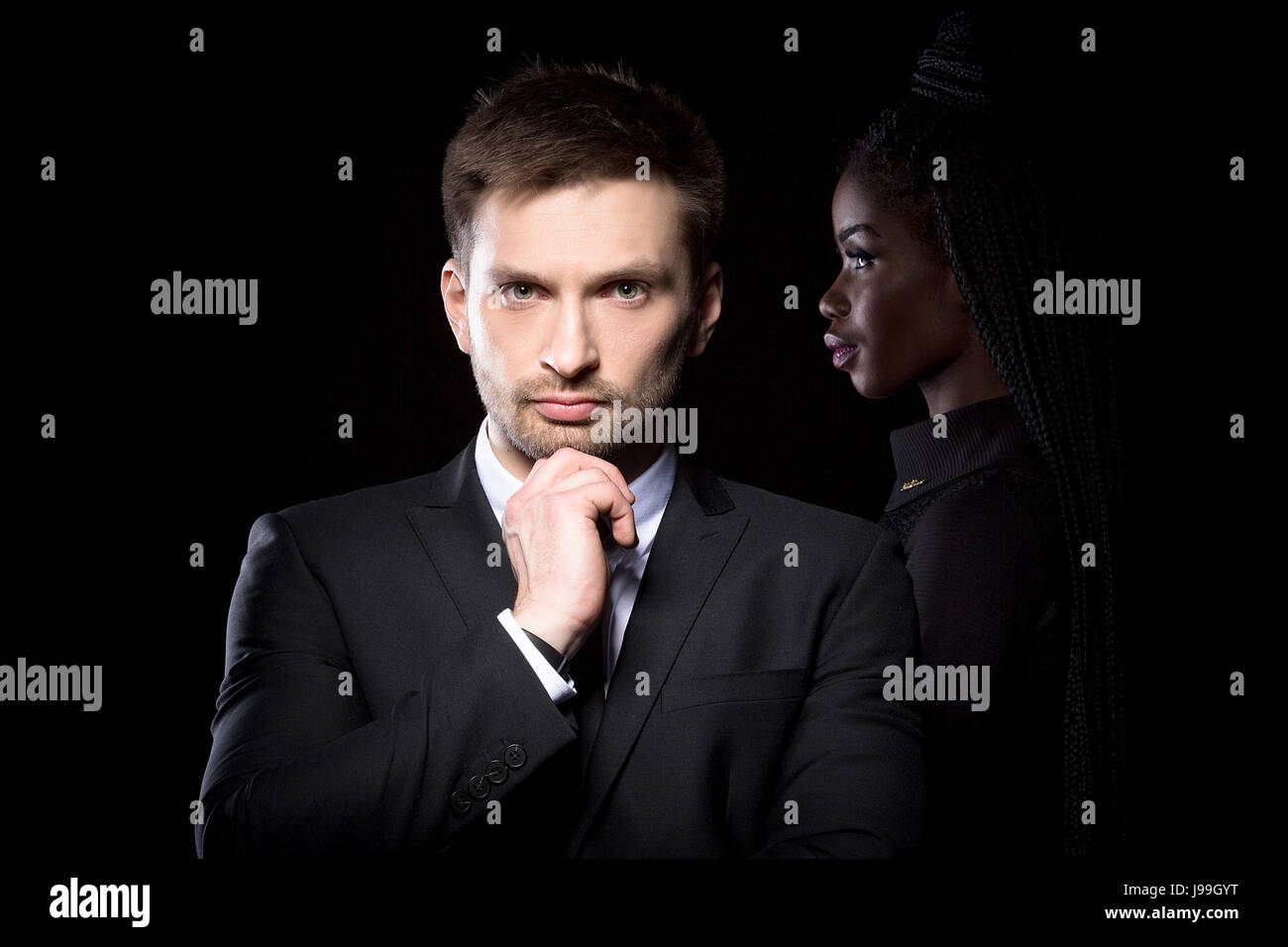 Man in suit standing with hand touching his chin with dark skinned woman behind. Stock Photo