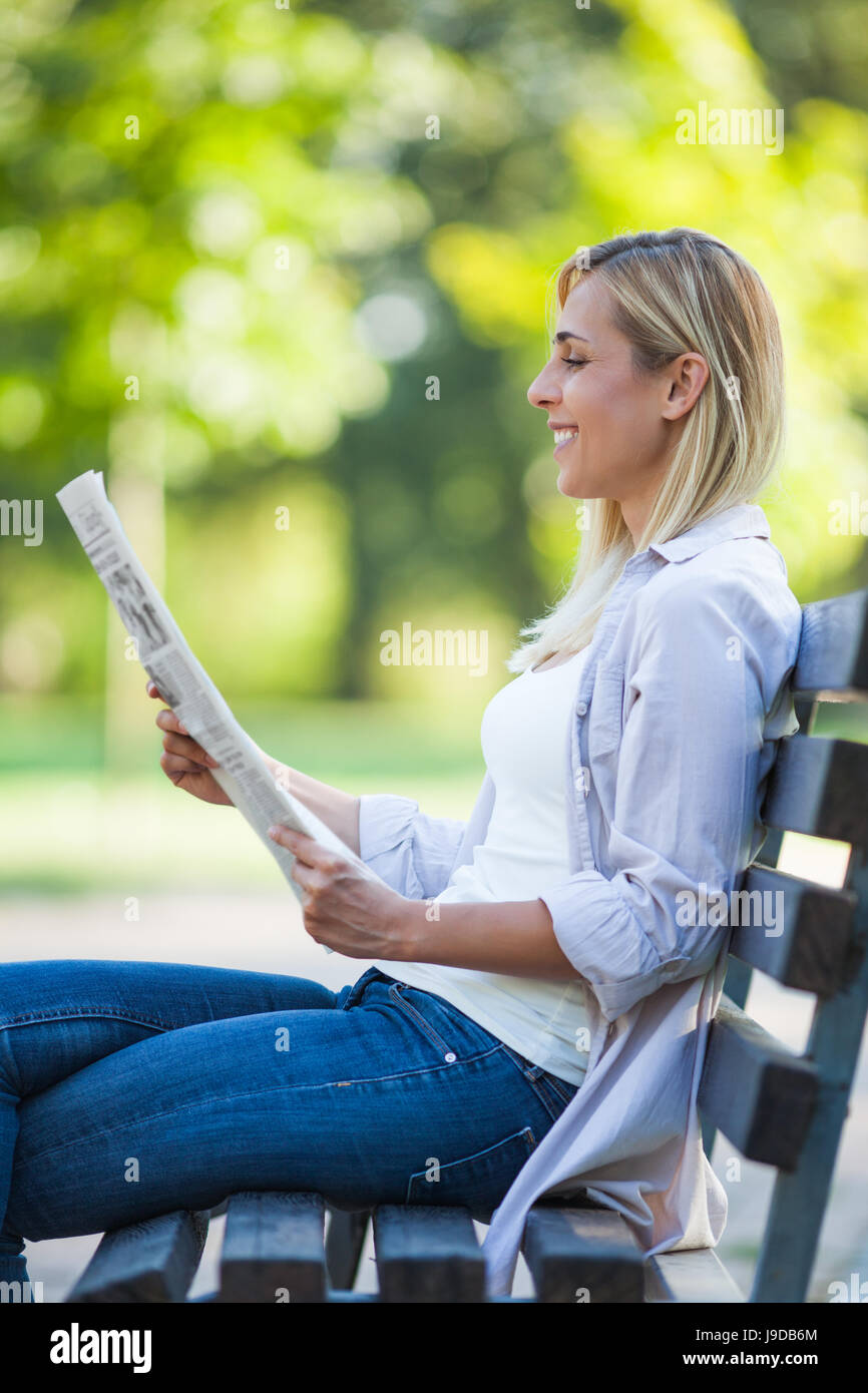 Young happy woman is sitting in park. She is reading newspapers. Stock Photo