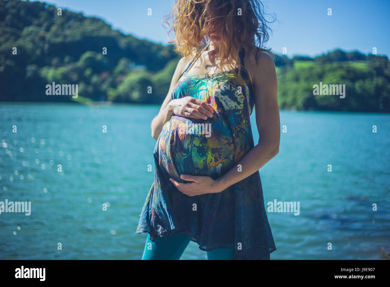 A young pregnant woman is standing by the river in a forest on a sunny