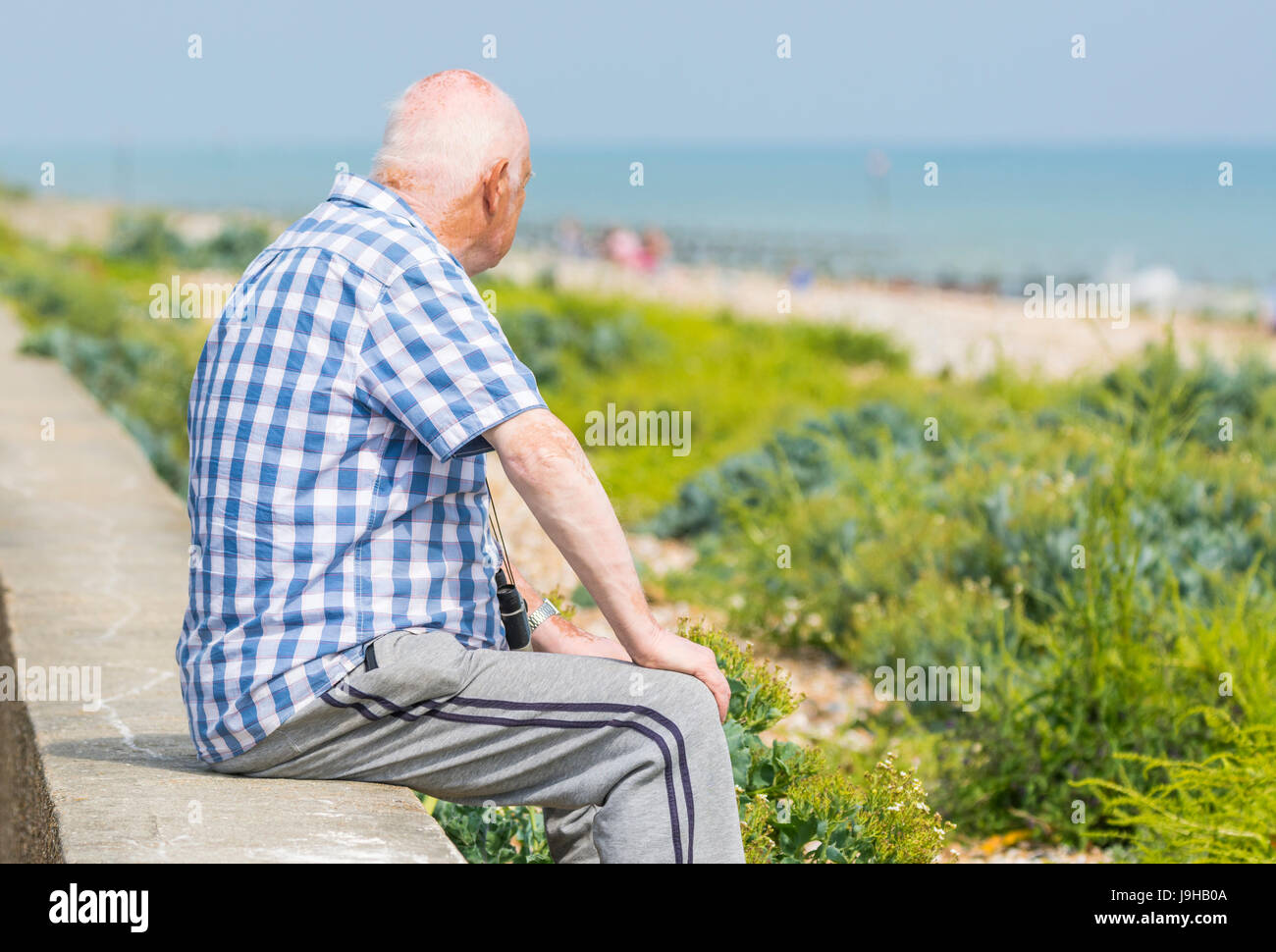 Elderly man sitting alone on a wall looking out to sea on a hot day in the UK. Stock Photo