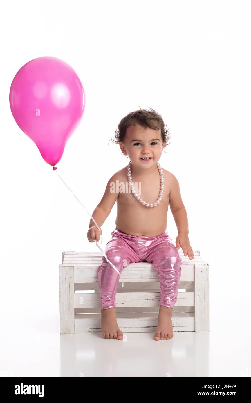 A happy, one year old, baby girl holding a pink balloon. She is wearing metallic pink leggings, a string of pearls and sitting on a wooden crate on a  Stock Photo