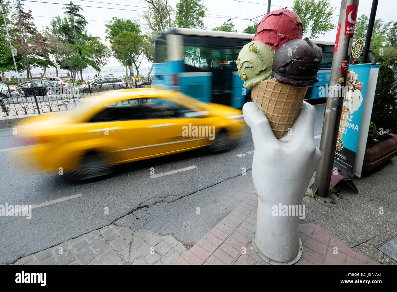 A giant hand holding an ice cream on a sidewalk in Istanbul, Turkey, with a yellow taxi and bus driving past. Stock Photo