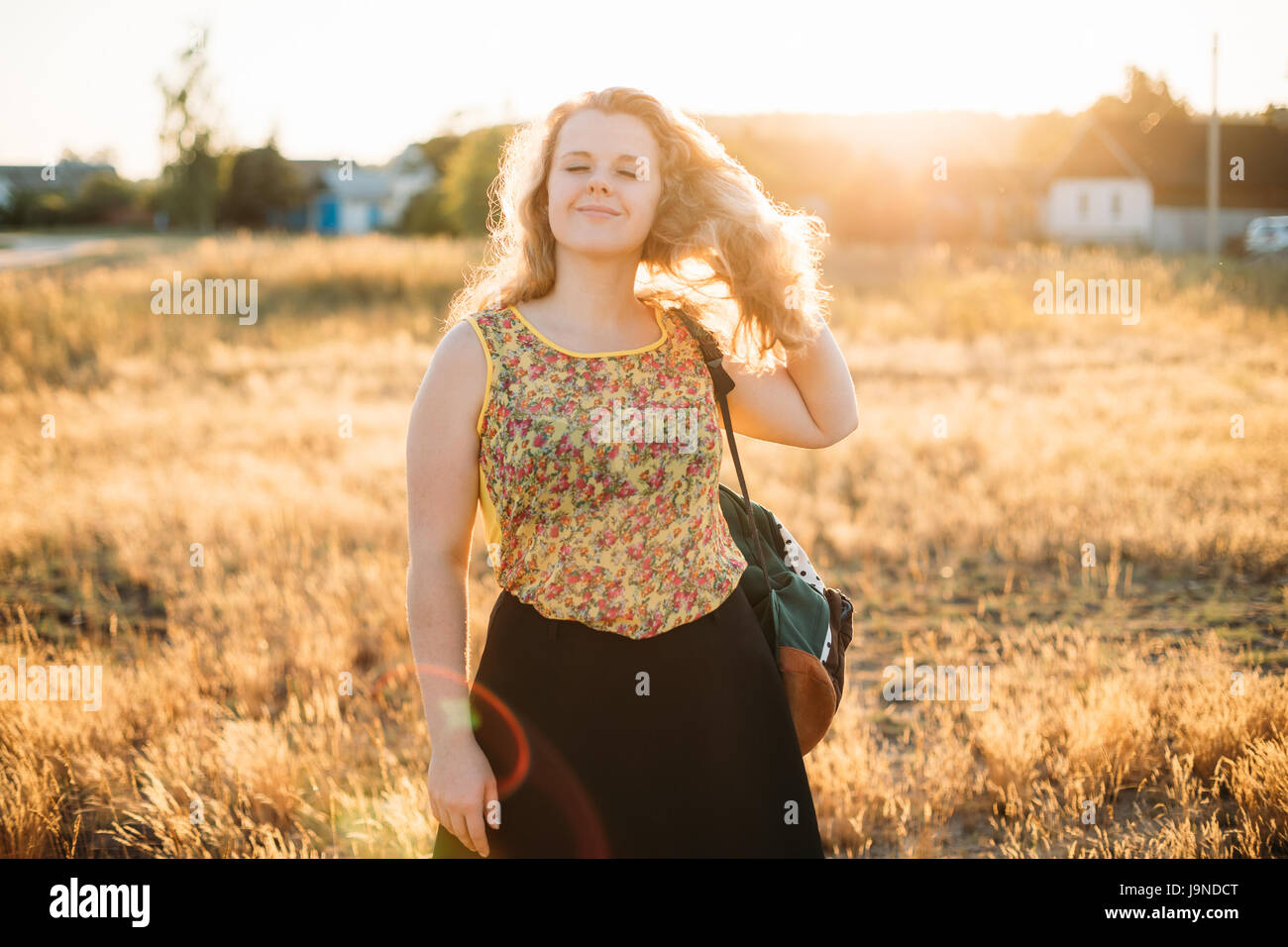 Portrait Of Young Pretty Plus Size Caucasian Happy Girl Woman With Closed Eyes, Wavy Brown Long Hair And Backpack In Summer Meadow Stock Photo