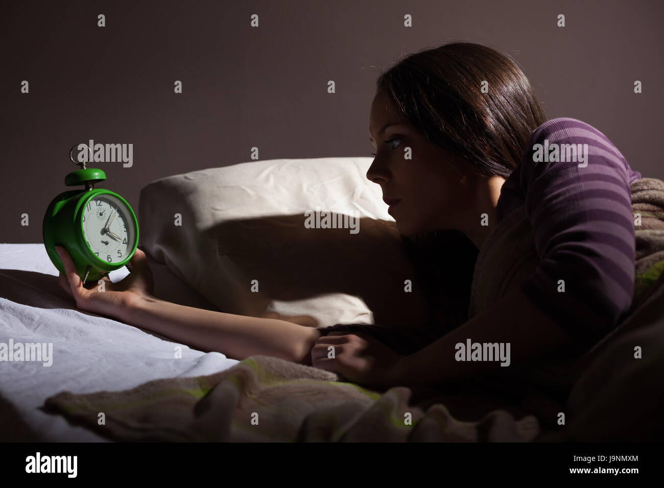 Young woman looking at clock at night. She has problems with sleep. Stock Photo