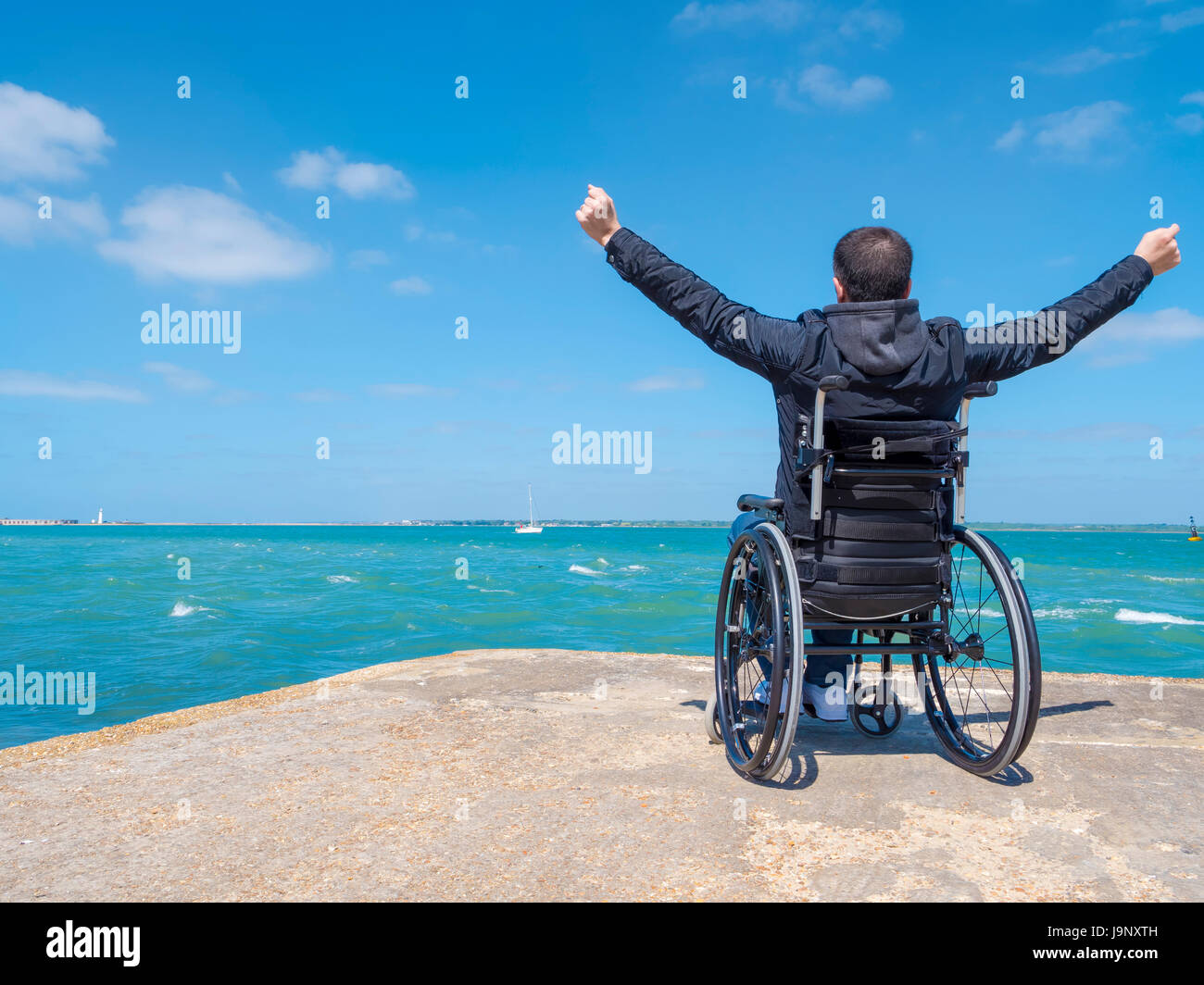 Disabled alone young man sitting in a wheelchair and looks at the sea. Stock Photo