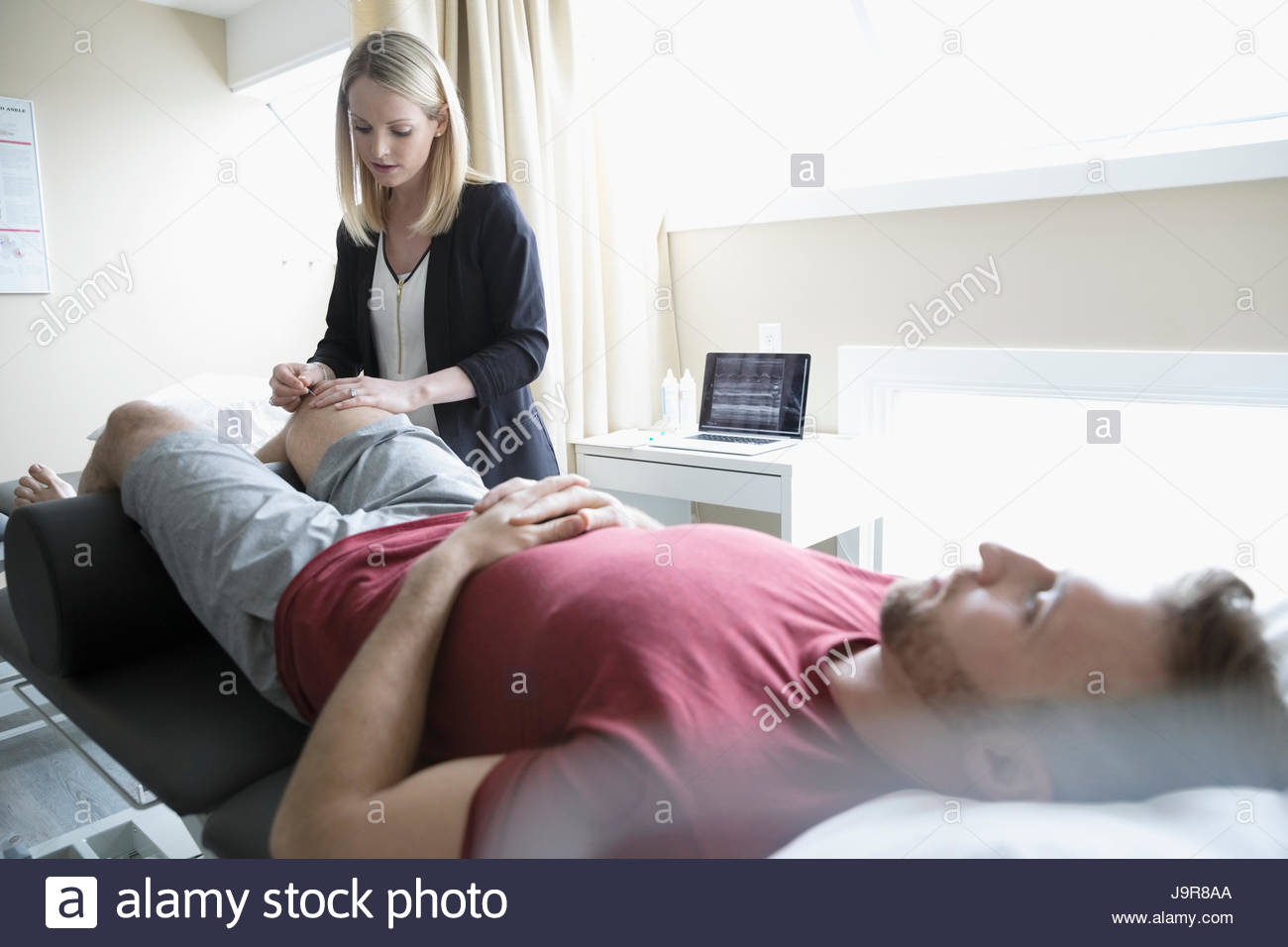 Female acupuncturist inserting needles into knee of man on clinic examination table Stock Photo