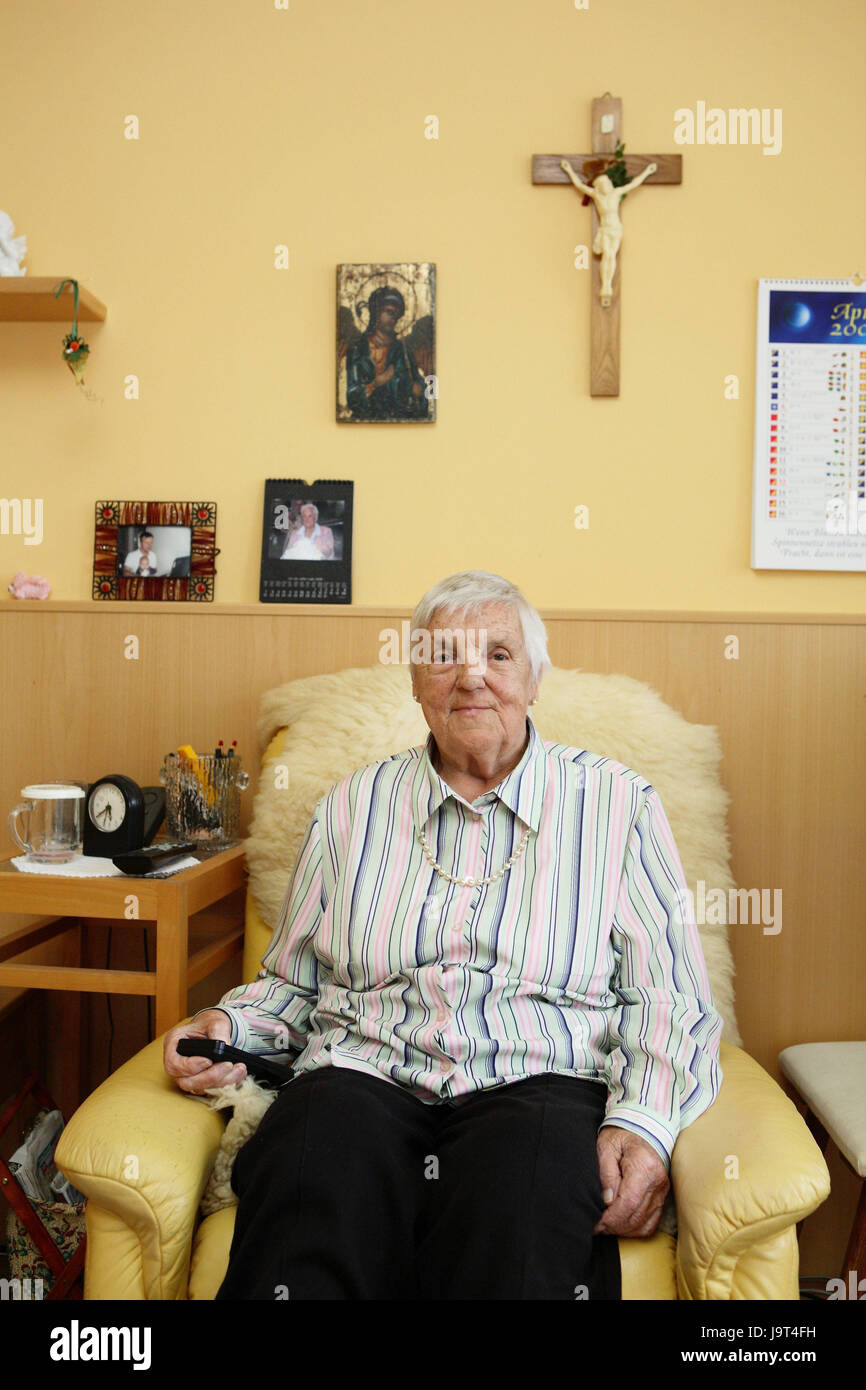 Old people's home,room,senior,armchair,sit,press remote control,people,old person,old,home inhabitant,nursing home,nursing home for the elderly,woman,old age,only,loneliness,pictures,cross,television armchair,watch TV,leisure time,activity,smile, Stock Photo