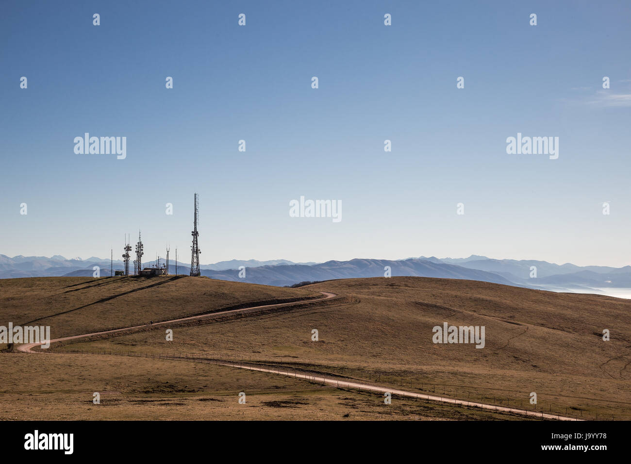 A curvy road towards some antennas on a top of mountain, under a blue, empty sky Stock Photo