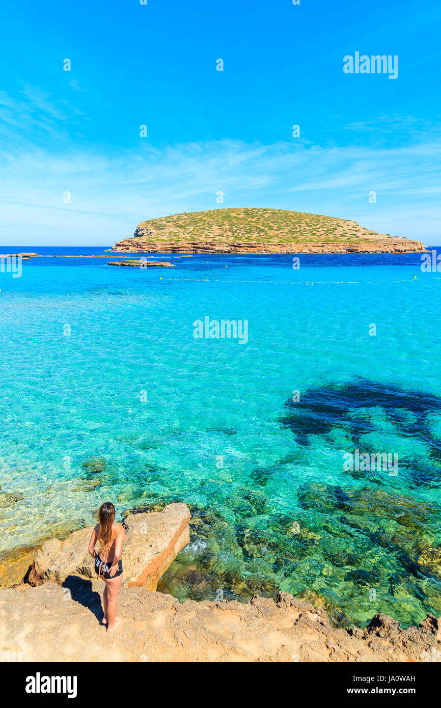 Unidentified young woman in swimsuit standing on a rock and posing against beautiful Cala Comte beach, Ibiza island, Spain Stock Photo