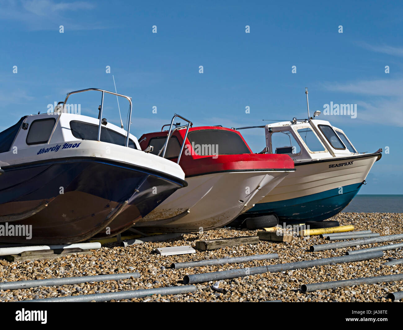 Row of three, small, sunlit boats beached on Eastbourne shingle with blue sky above, Eastbourne, East Sussex, England, UK Stock Photo