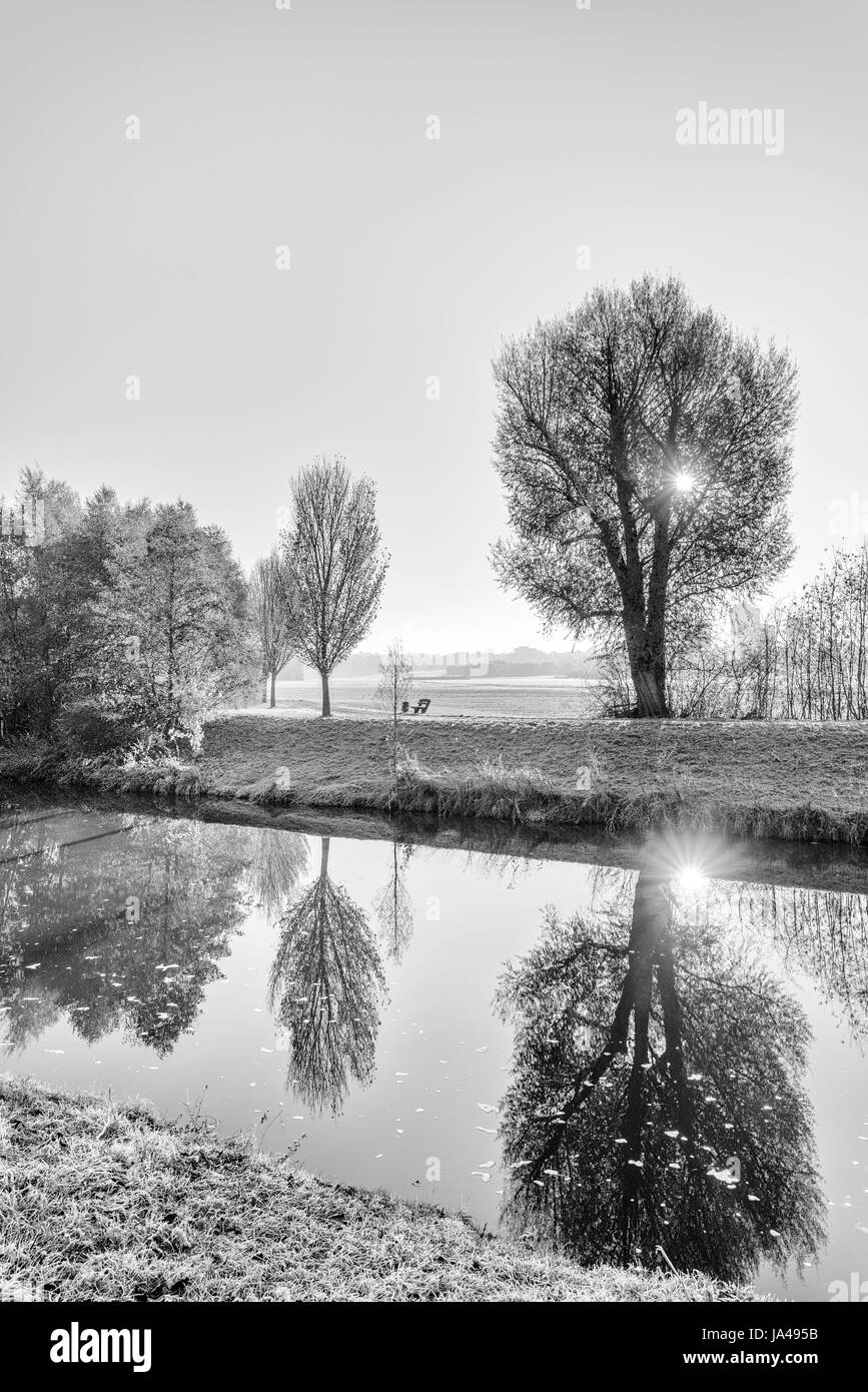 Monochrome high key scenic photo of an autumnal idyllic sunrise at a river with water reflections of trees and the sun,sunrays,path,wanderlust Stock Photo