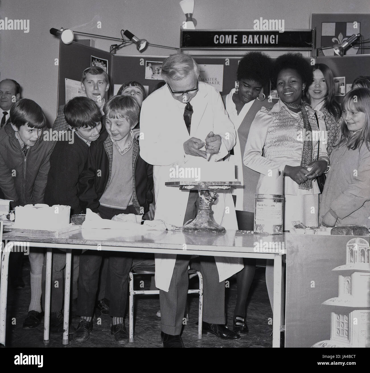 1960s, historical, come baking!.. a group of school pupils at a careers show, watch a man in a white coat icing a cake on a baking stand, South-east London, England, UK. In this era, both boys and girls attended classes in what was known as 'home economics', which covered areas such as heathly foods, cooking and household budgeting. Stock Photo