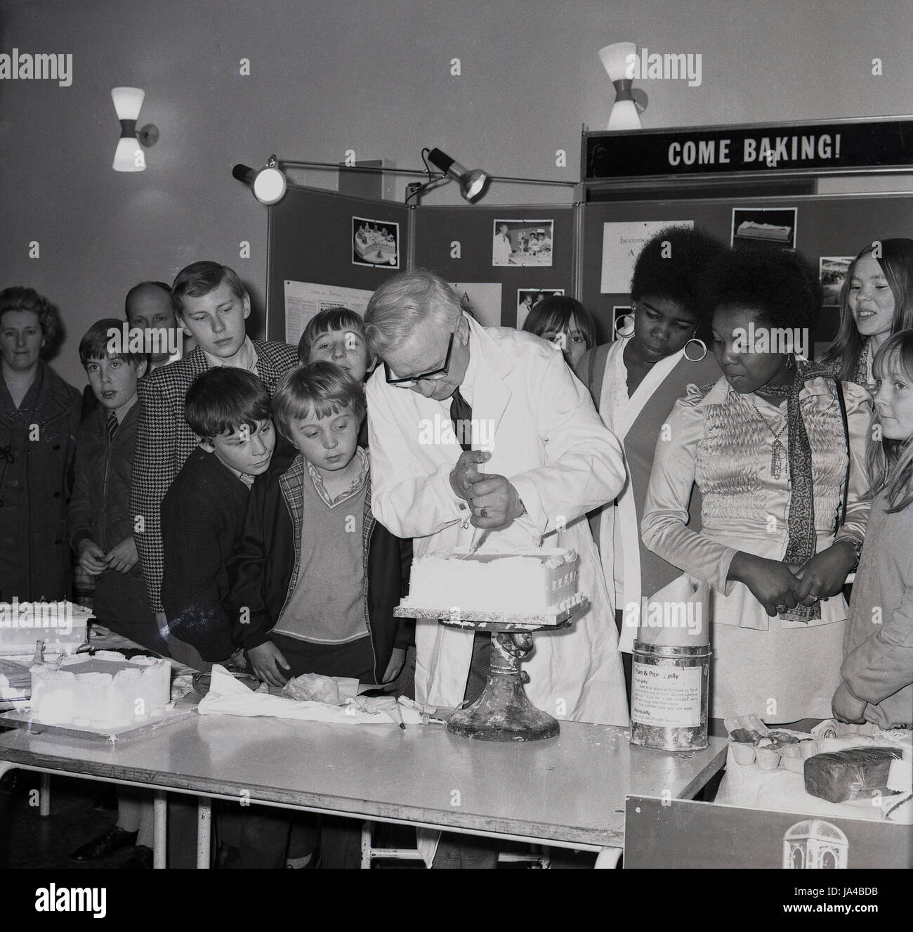 1960s, historical, come baking!.. a group of school pupils at a careers show, watch a man in a white coat icing a cake on a baking stand, South-east London, England, UK. In this era, both boys and girls attended classes in what was known as 'home economics', which covered areas such as foods, cooking and household budgeting. Stock Photo