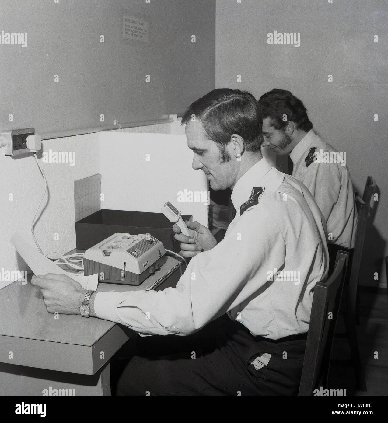 1970s, two uniformed Police officers sitting at booths using the new dictaphone or audio recording machines the Metropolitan Police service introduced in this era to file a suspect's statements or crime reports. Stock Photo