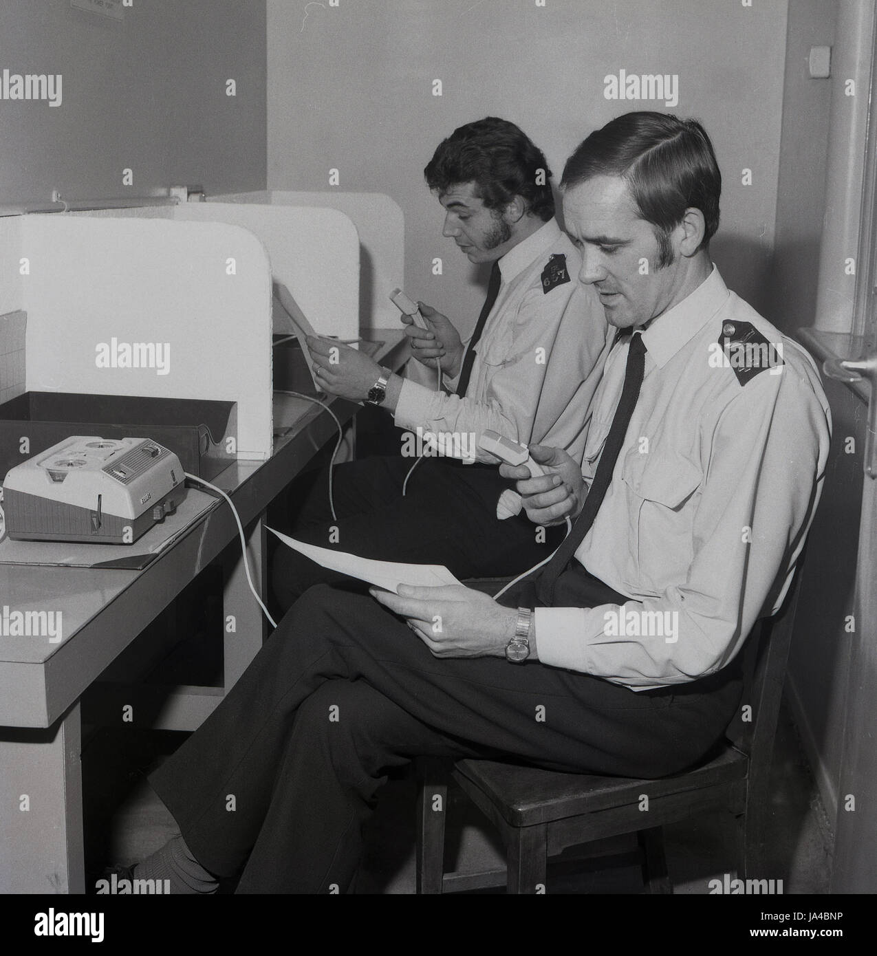 1970s, two uniformed Police officers sitting at booths using the new dictaphone or audio recording machines the Metropolitan Police service has introduced to to file statements or reports. Stock Photo