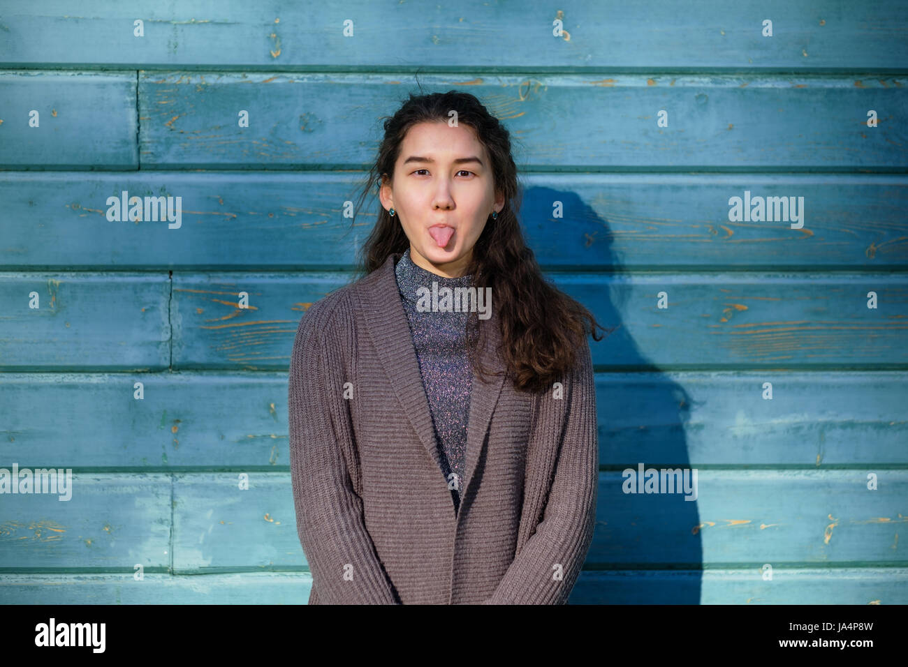 A beautiful Asian girl fools around and shows her tongue. She stands by the wooden blue wall. She wears a warm jacket. Stock Photo