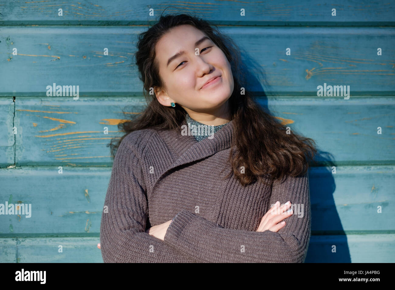 A beautiful Asian girl with long hair and a warm sweater is standing by the wooden blue wall. She looks forward with smile Stock Photo