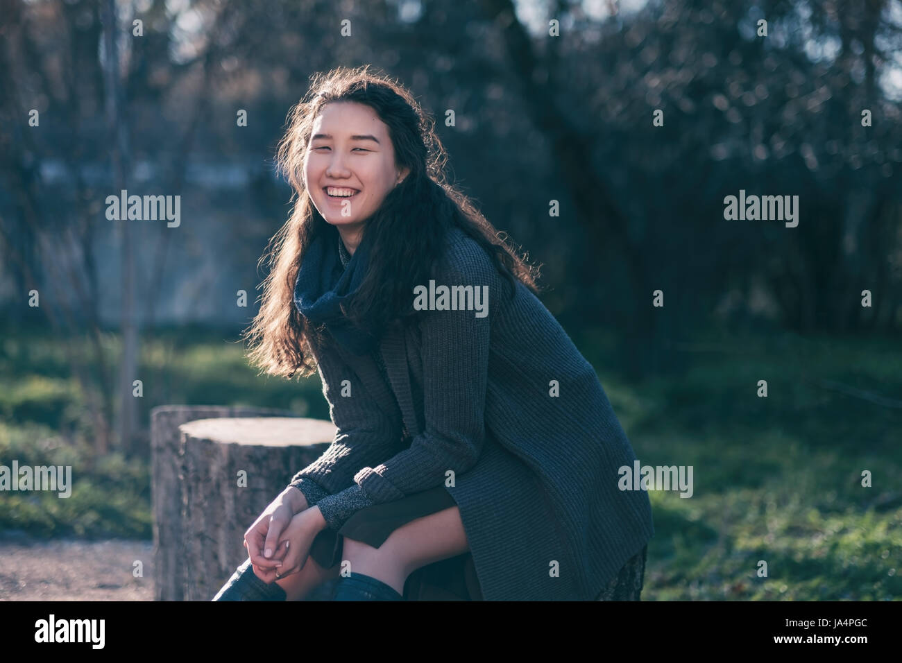 A beautiful Asian girl sits in a park on a stump and smiles. Enjoy the silence in city life. Stock Photo