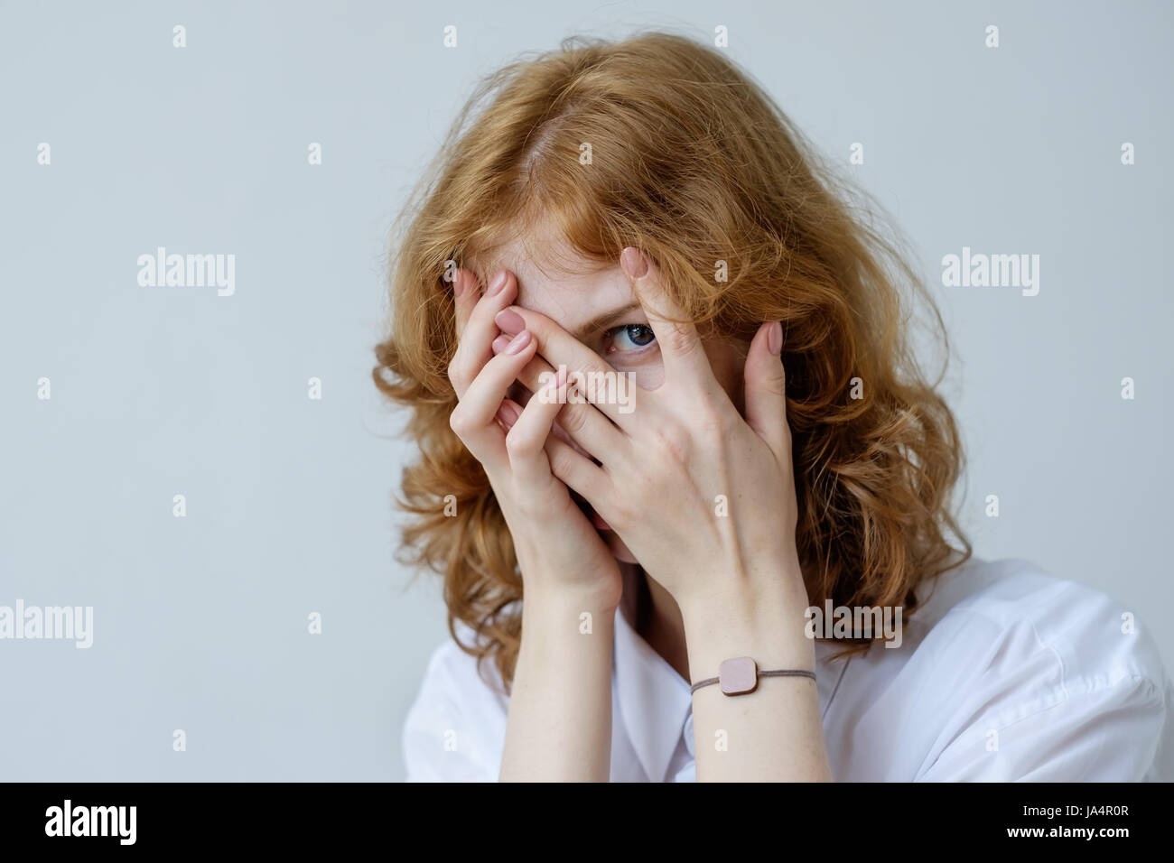 A red-haired girl with curls in embarrassment covers her face with her hands and looks through her fingers. Social phobia of the younger generation. Stock Photo