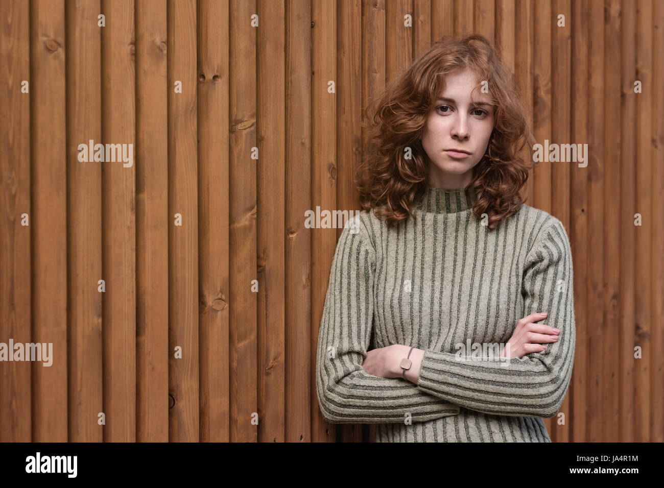 A beautiful red-haired girl in a green sweater is standing by the wooden wall. She crosses her arms and looks severely at the camera. Stock Photo