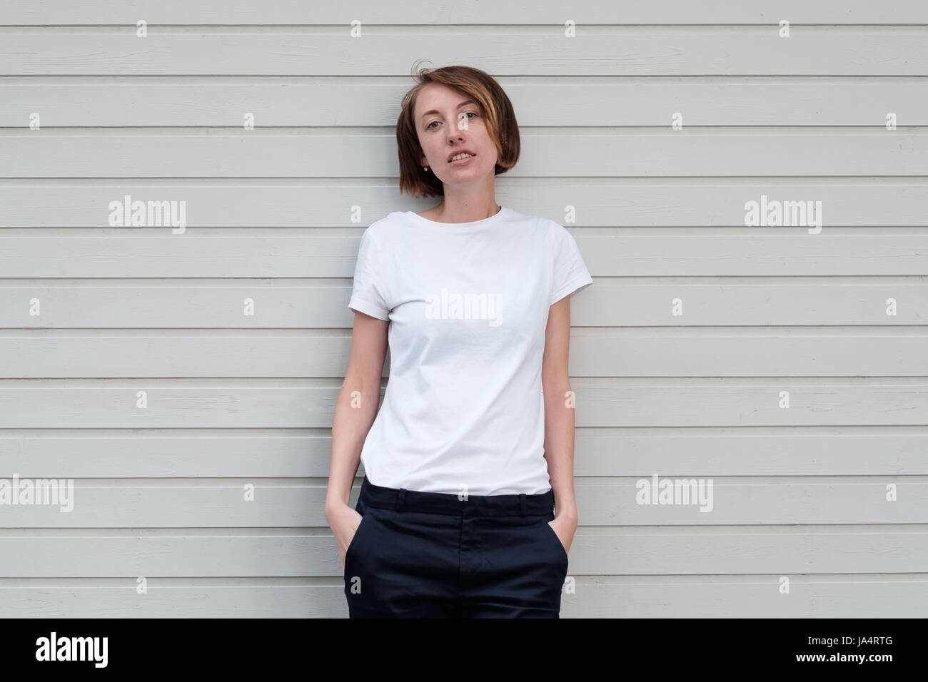 A beautiful girl with freckles in a white T-shirt is standing by the wooden wall. Stock Photo