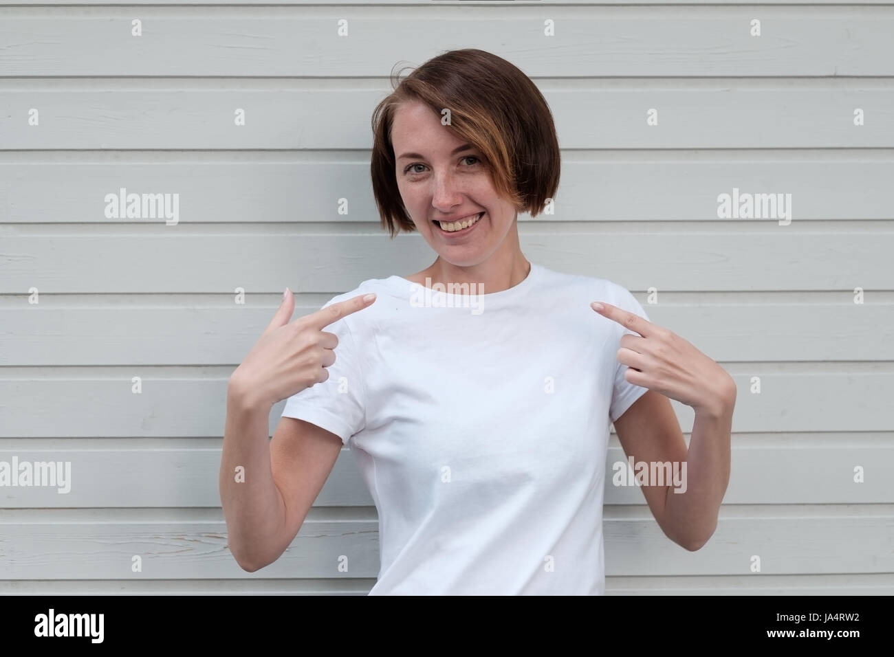 A beautiful girl with freckles in a white T-shirt is standing by the wooden wall. She points to herself with her fingers, clarifying the choice Stock Photo