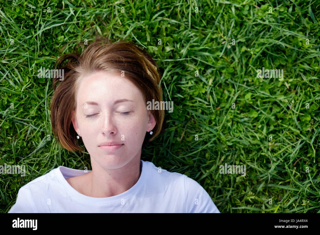 A beautiful girl with freckles lies on the green grass with her eyes closed and rests Stock Photo