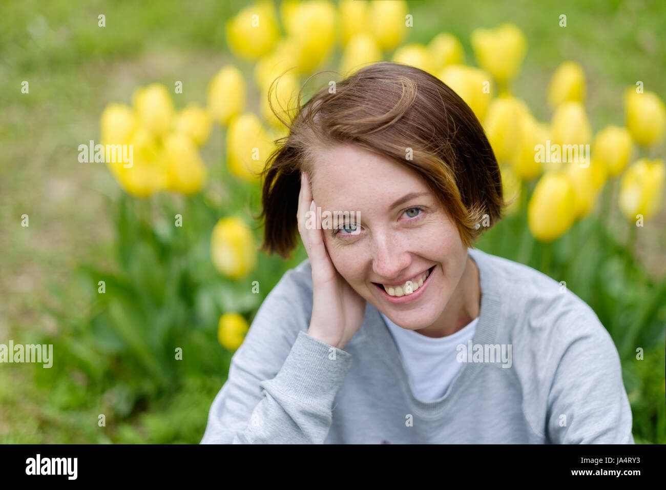 A girl with freckles sits on the grass beside the yellow tulips in the park. Stock Photo