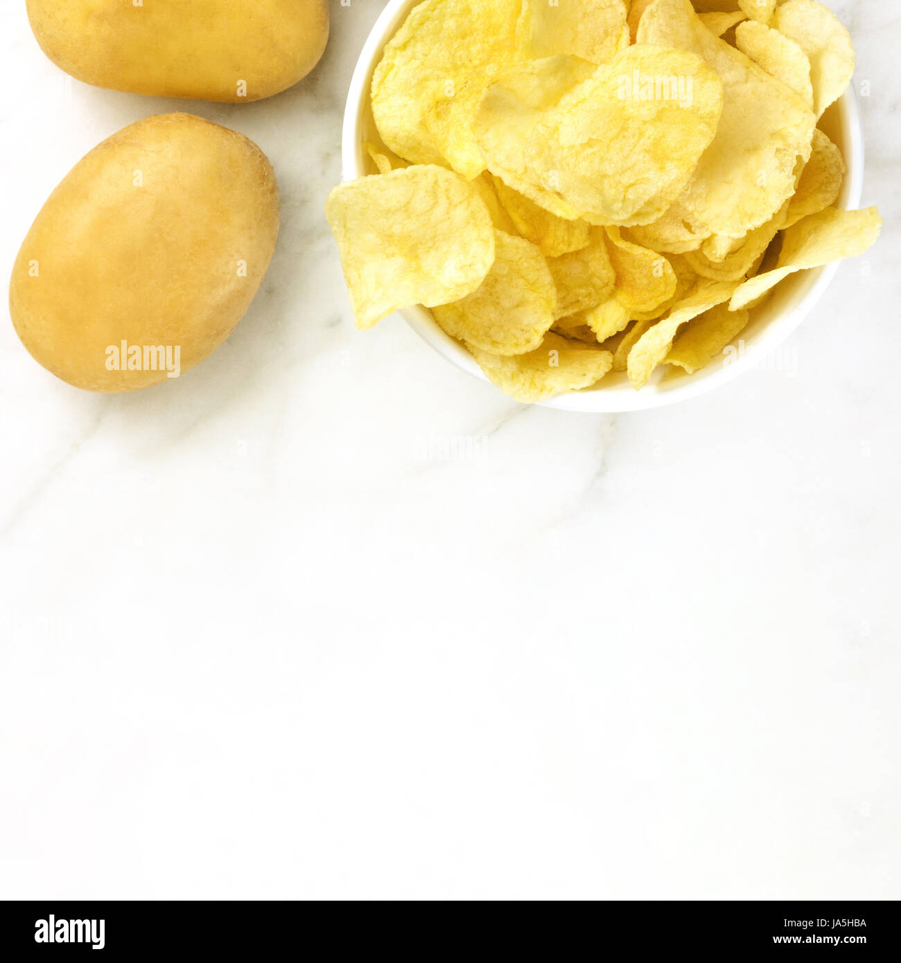 A square photo of a bowl of potato chips with potatoes, shot from above on a white marble texture with a place for text Stock Photo