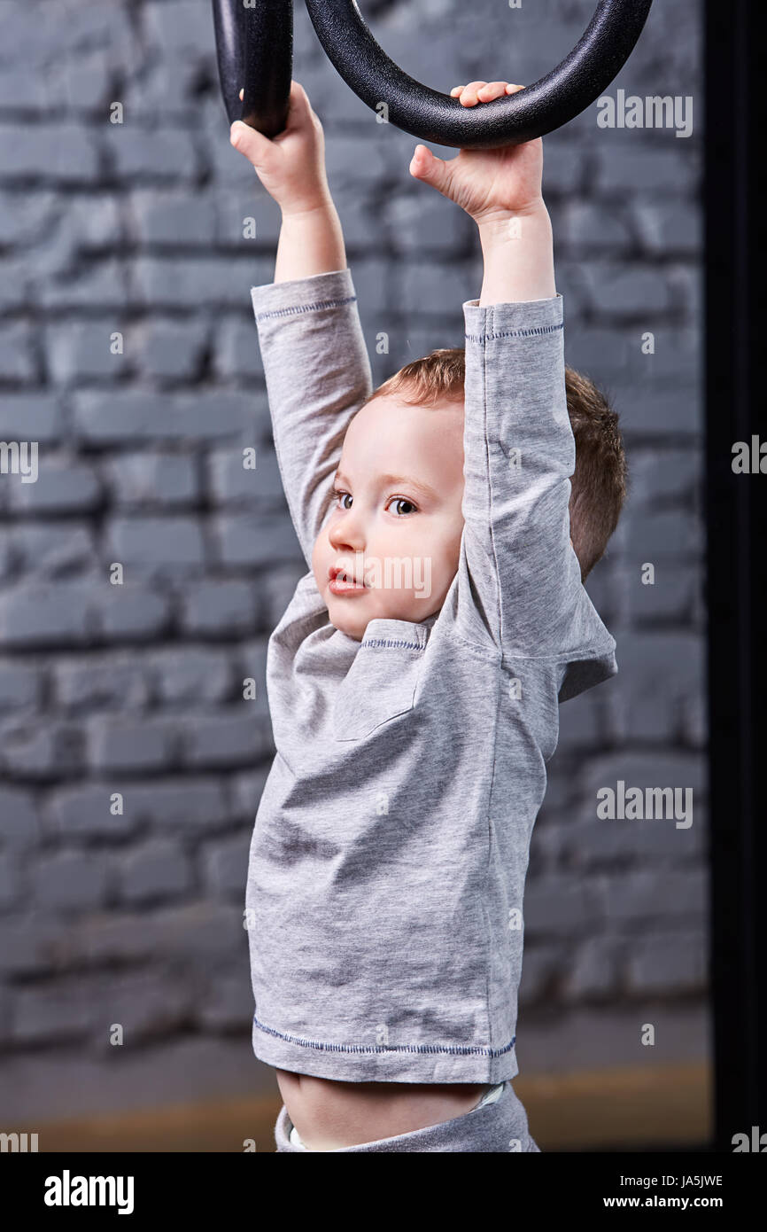 Little child boy in tge grey sportwear hanging on gymnastic rings