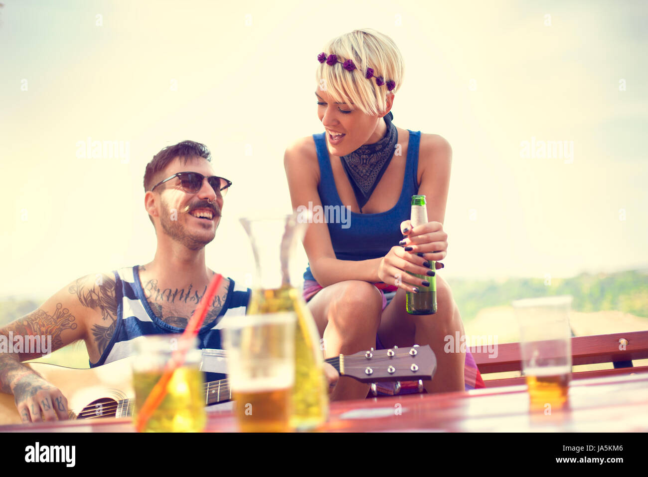 Heavily tattooed guy playing guitar while lassies drinks beer in nature at spring Stock Photo