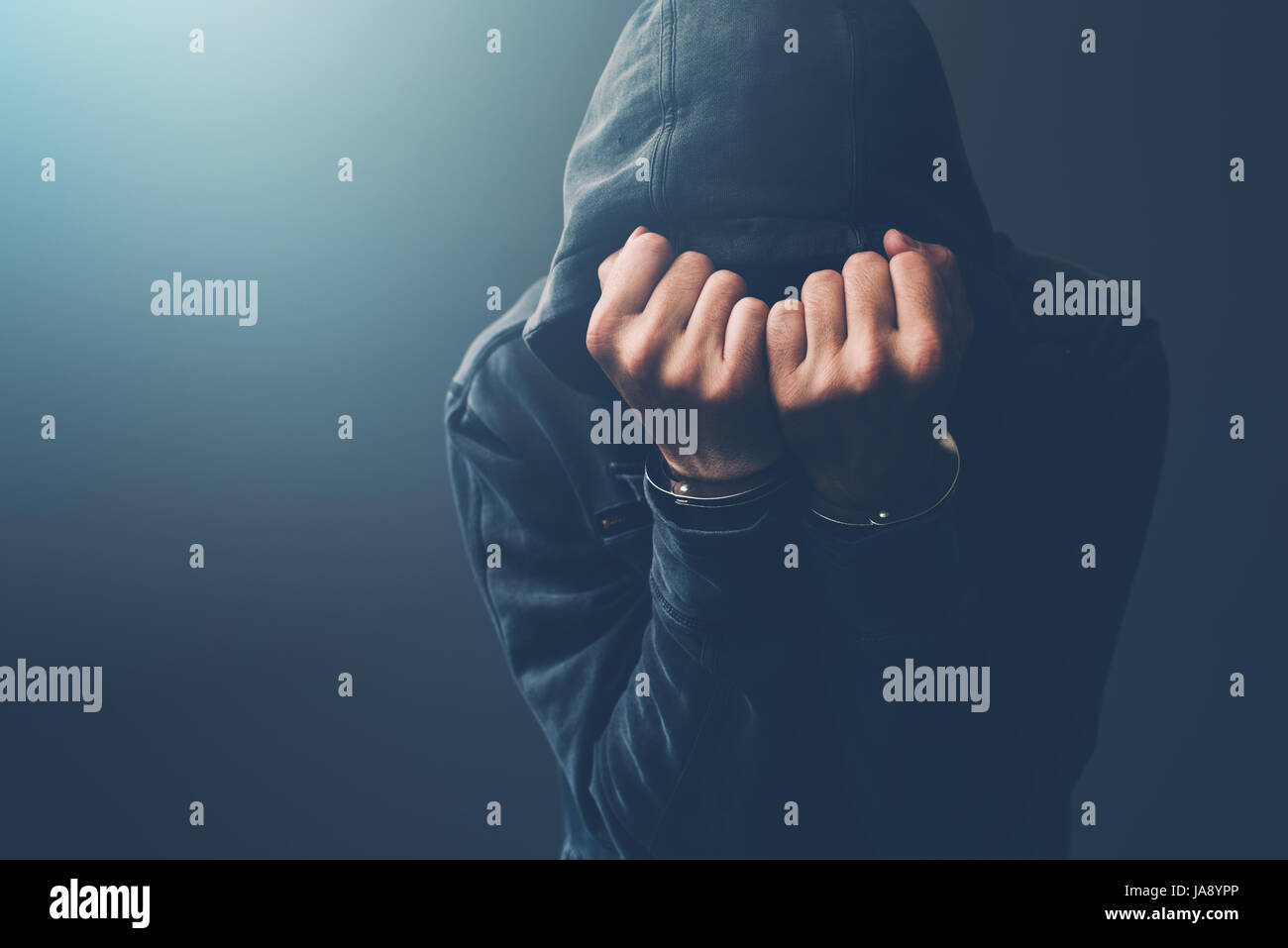 Arrested computer hacker and cyber criminal with handcuffs wearing hooded jacket hiding face Stock Photo