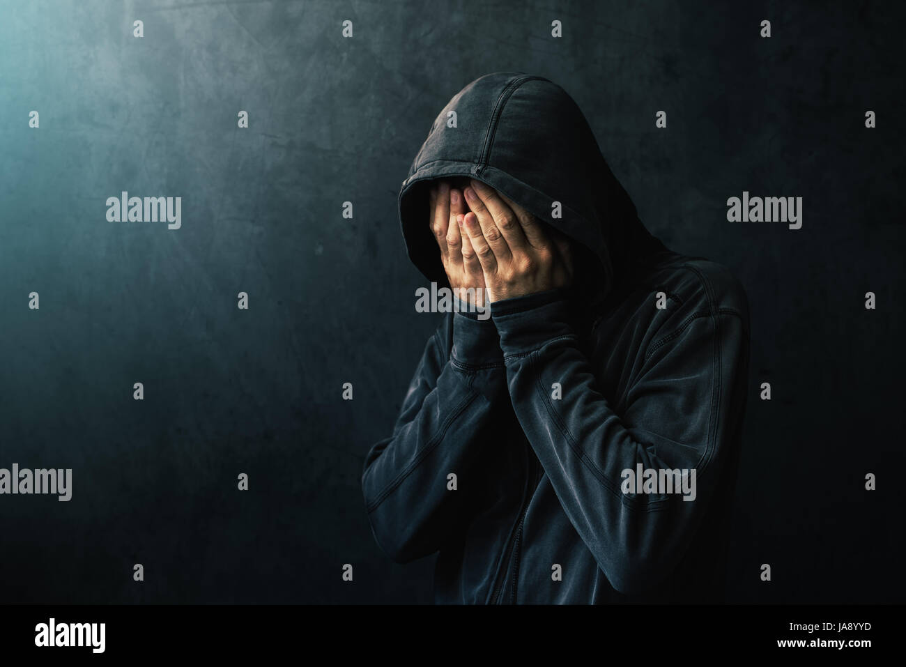 Desperate man in hooded jacket is crying, hands are covering face and tears in the eyes, light of hope shining from his right side Stock Photo