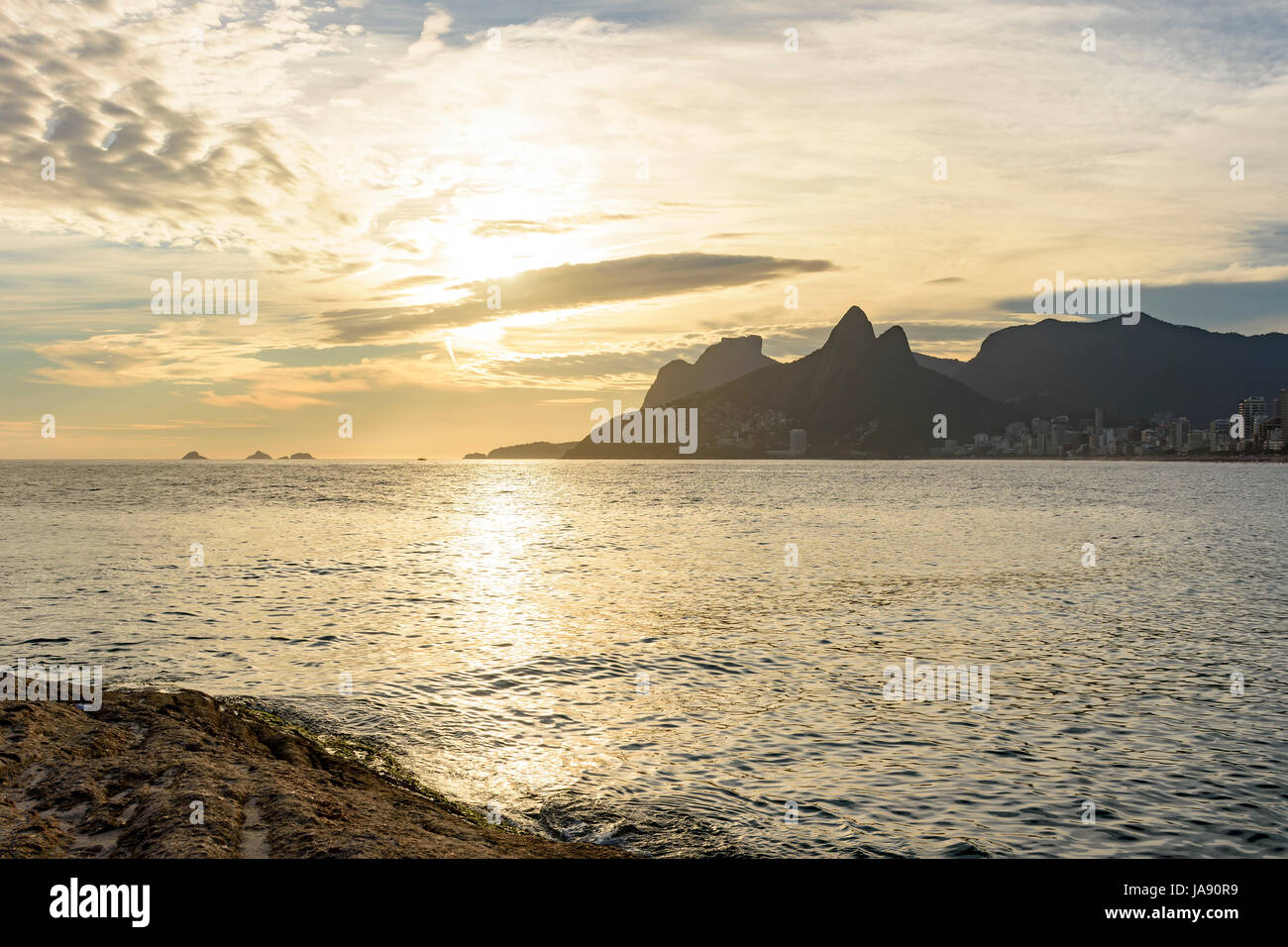 Arpoador beach in Rio de Janeiro, with its stones, buildings and the seaside during sunset and the Two Brothers hill in background Stock Photo