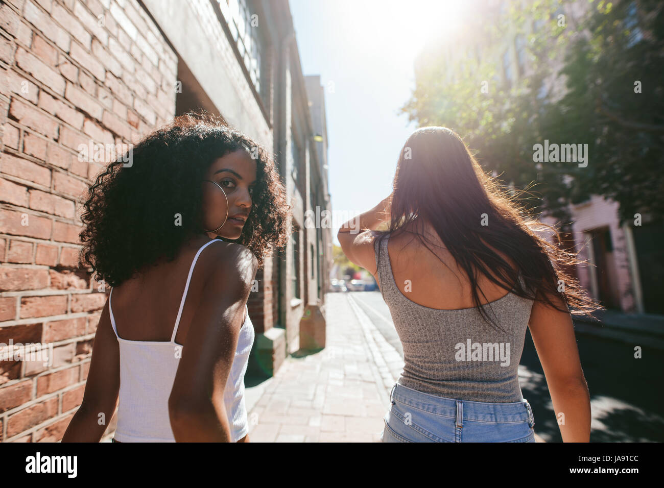 Rear view shot of young african woman looking over her shoulder while walking with her friend on city street. Two young women walking together outdoor Stock Photo