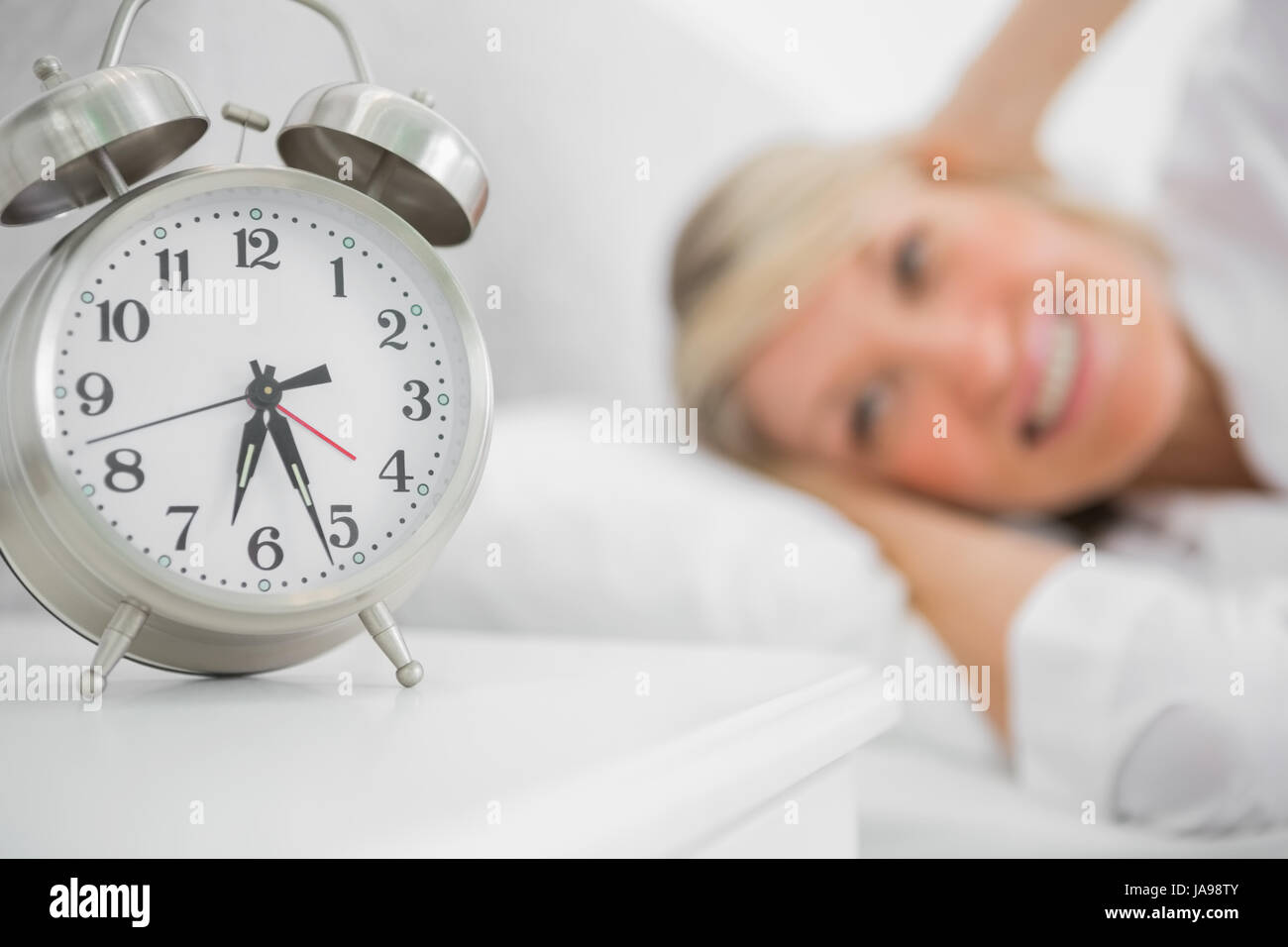 Alarm clock annoying blonde woman in bed blocking her ears Stock Photo