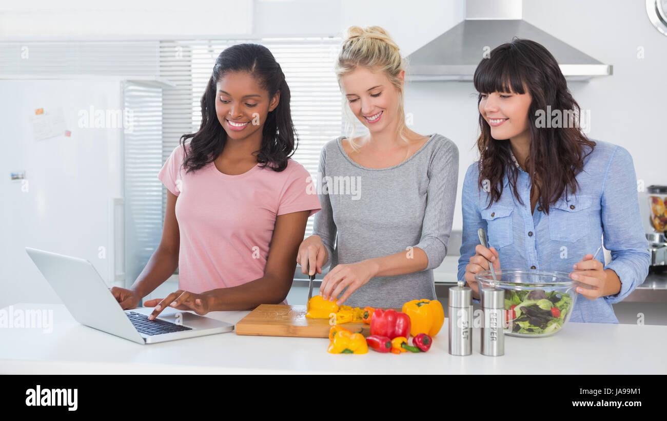Happy friends making salad and using laptop for recipe at home in kitchen Stock Photo