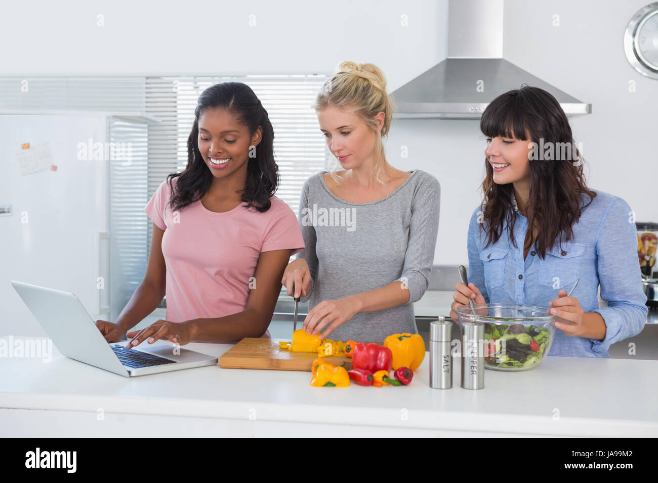 Smiling friends making salad and using laptop for recipe at home in kitchen Stock Photo