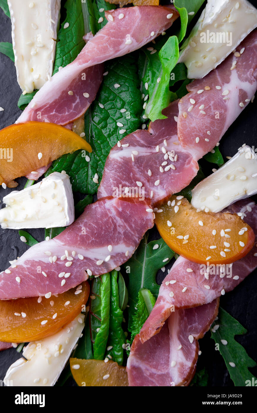 Salad with ham jamon serrano, camembert, melon, arugula on black stone slate plate on black background. Top view. Stock Photo