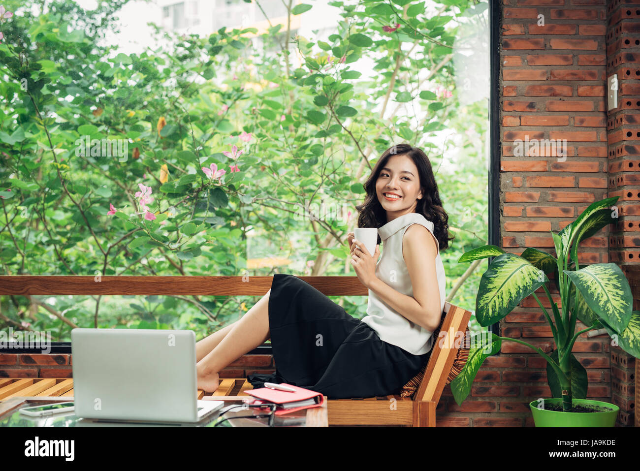 Confident young asian woman in smart casual wear holding coffee mug while sitting near window in creative office or cafe Stock Photo