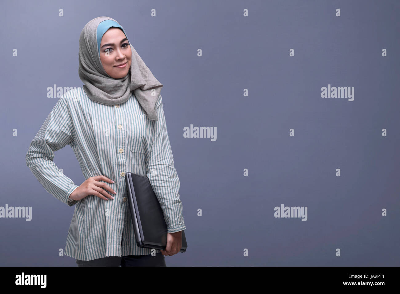 Happy asian muslim woman wearing veil holding laptop against dark ...