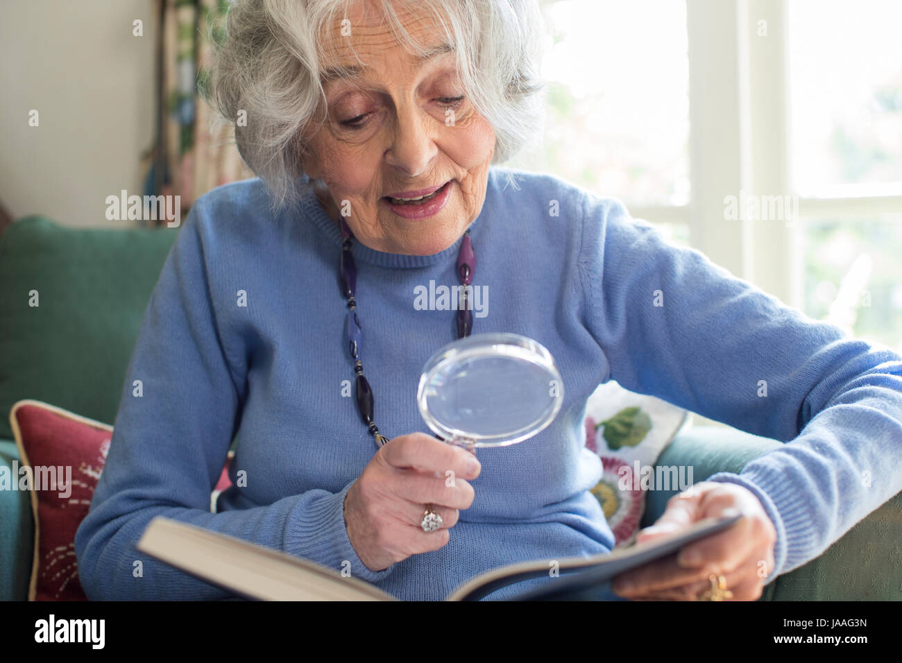 Senior Woman At Home Reading Book Using Magnifying Glass Stock Photo
