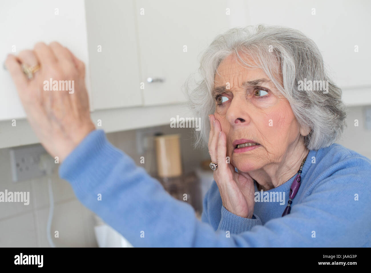 Forgetful Senior Woman With Dementia Looking In Cupboard Stock Photo