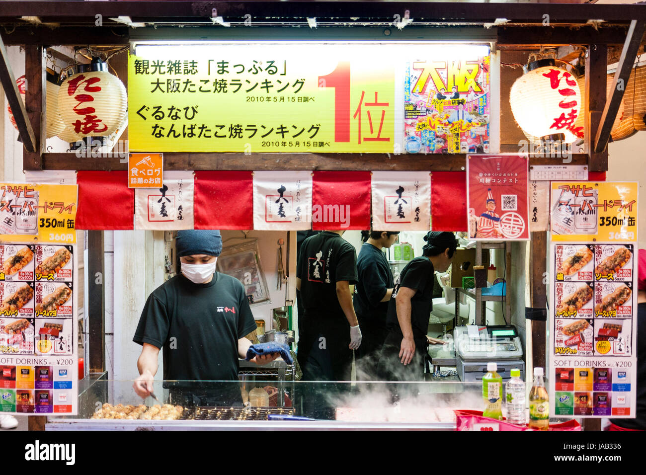 Small take-away hot food counter with cook, wears face mask, making Takoyaki, Octopus balls. Menu boards on both sides. Night time, Dotonbori, Osaka. Stock Photo