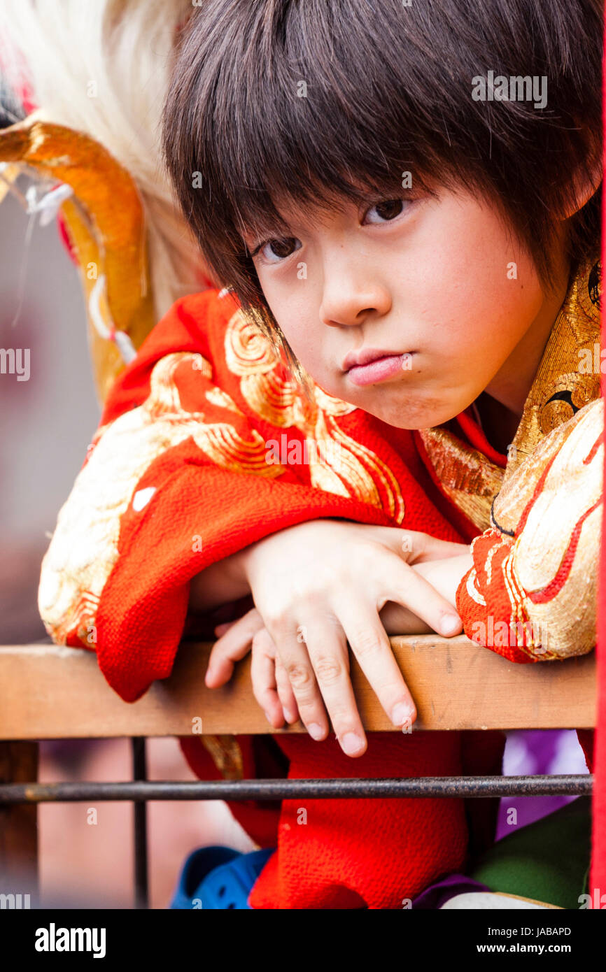 Japanese child, boy, 8-9 years old, leaning over wooden rail looking directly at viewer, unhappy, angry expression, eye-contact. Wears red yukata coat. Stock Photo