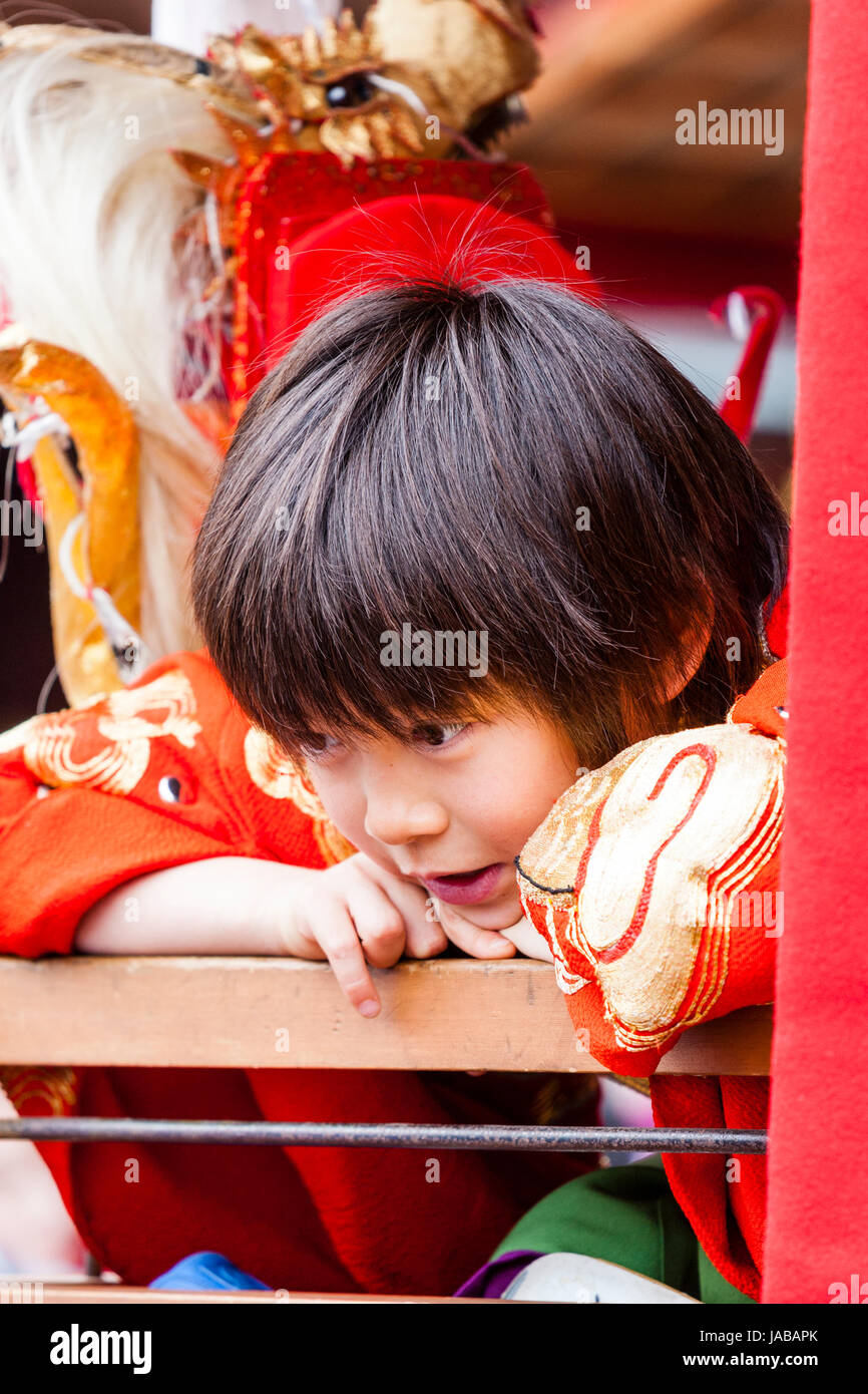 Japanese child, boy, 8-9 years old, leaning over wooden rail as he talks to someone (unseen) Wears a red and golden yukata coat. Stock Photo