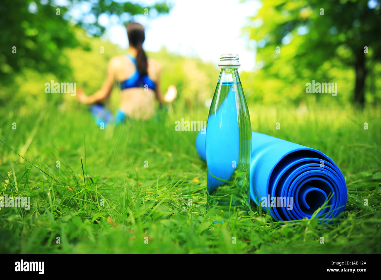 Healthy lifestyle background. Fitness theme. Fitness mat and bottle of water in green grass. Girl in yoga pose on background. Stock Photo