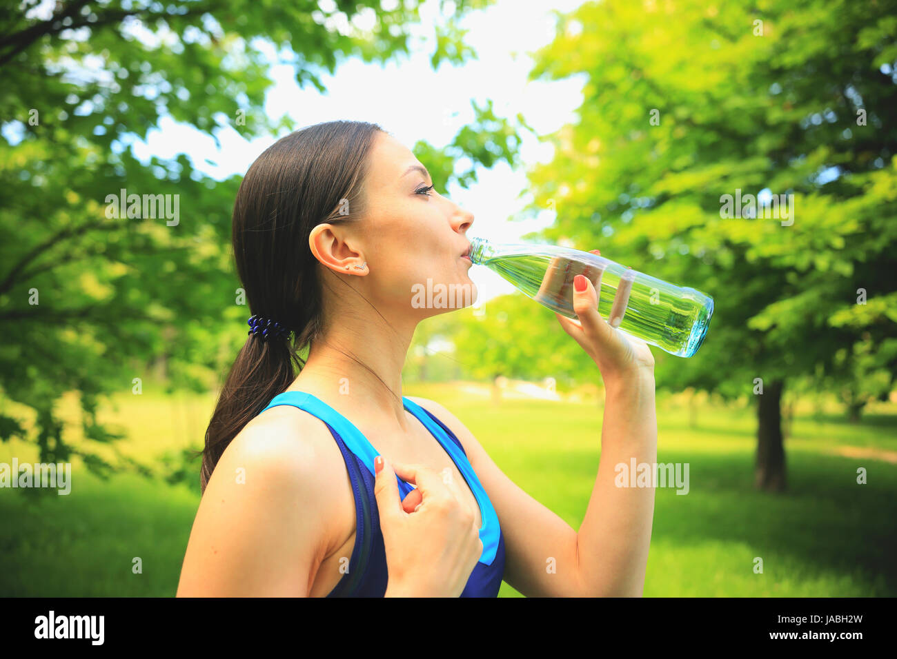 Sports outdoors scene. Brunette girl drinks water in park after fitness trainiing. Vivid sport background. Stock Photo
